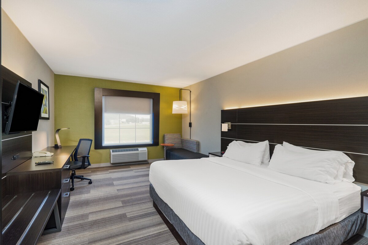 A hotel room with a large bed, a desk with a chair, a television, a window, and a wall-mounted lamp.