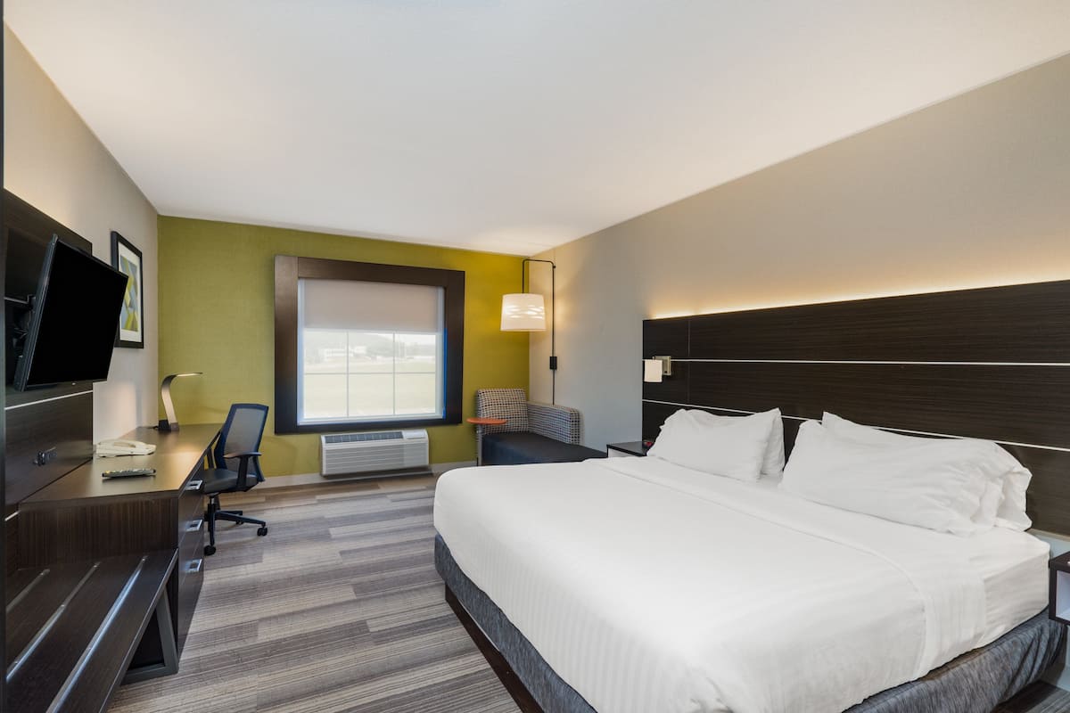 Habitación de hotel con una cama grande, un escritorio con silla, televisor, ventana y una lámpara de pared.