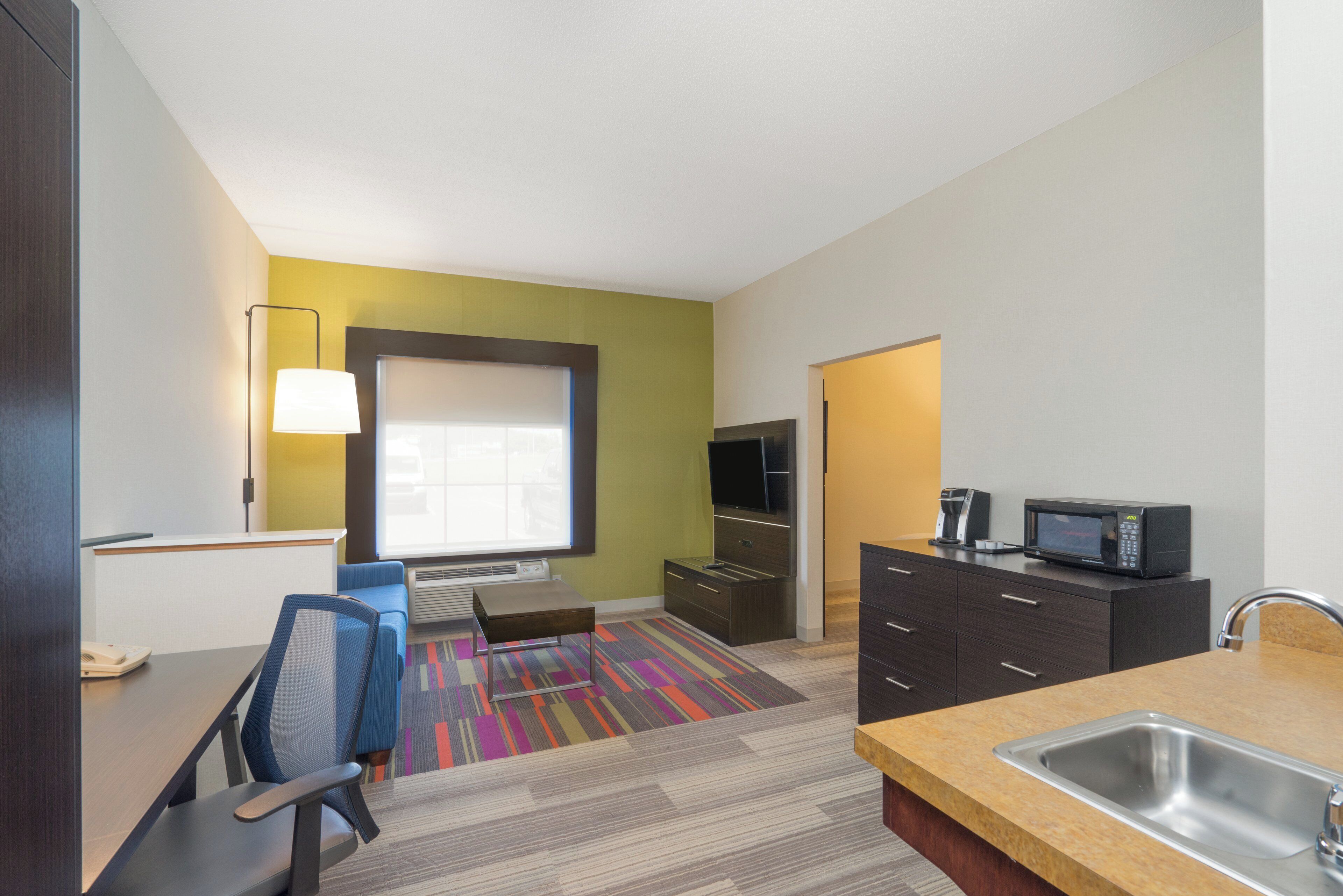 A hotel room with a kitchenette, a flat-screen TV, a sofa, a desk, and a chair.