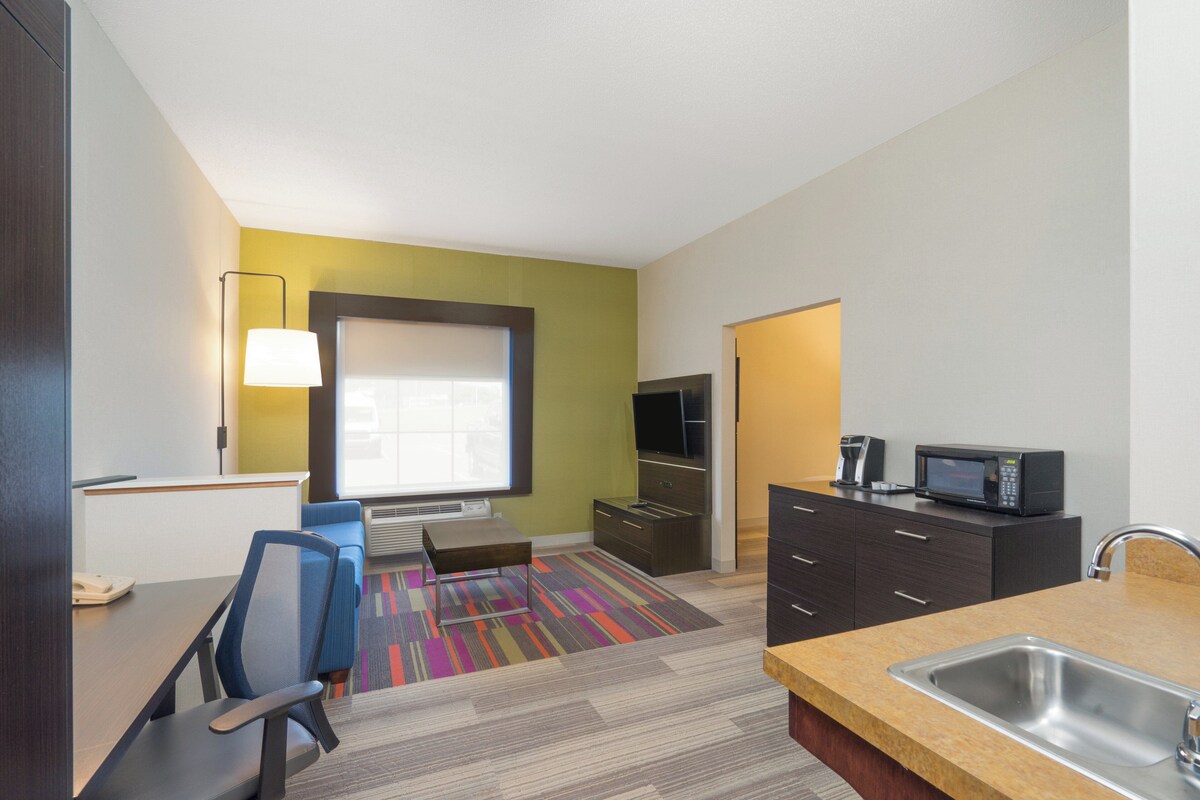 A modern hotel room with a kitchenette, a large window, a colorful rug, and a seating area with a chair and a small table.