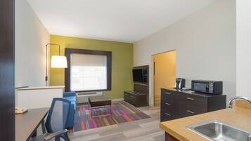 A hotel room with a kitchenette, a flat-screen TV, a sofa, a desk, and a chair.