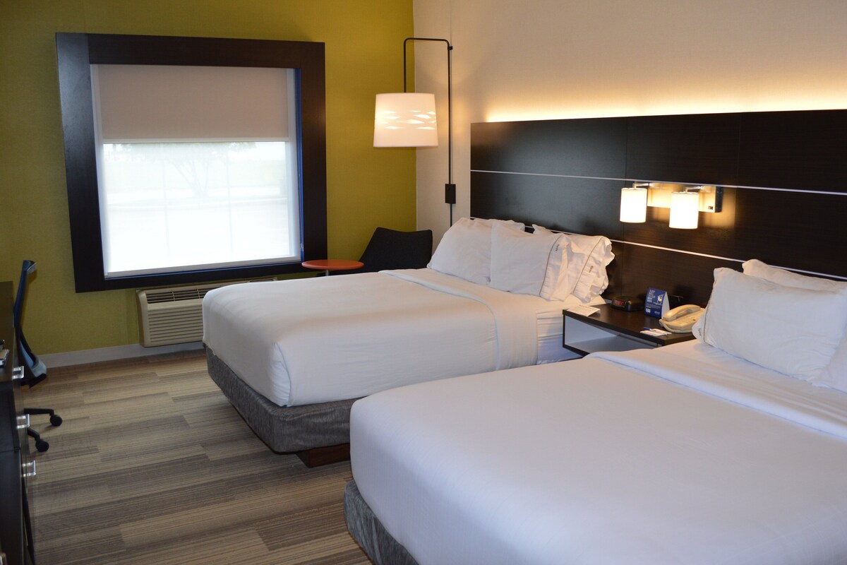 A hotel room with two beds, a desk, a chair, a lamp, and a window.