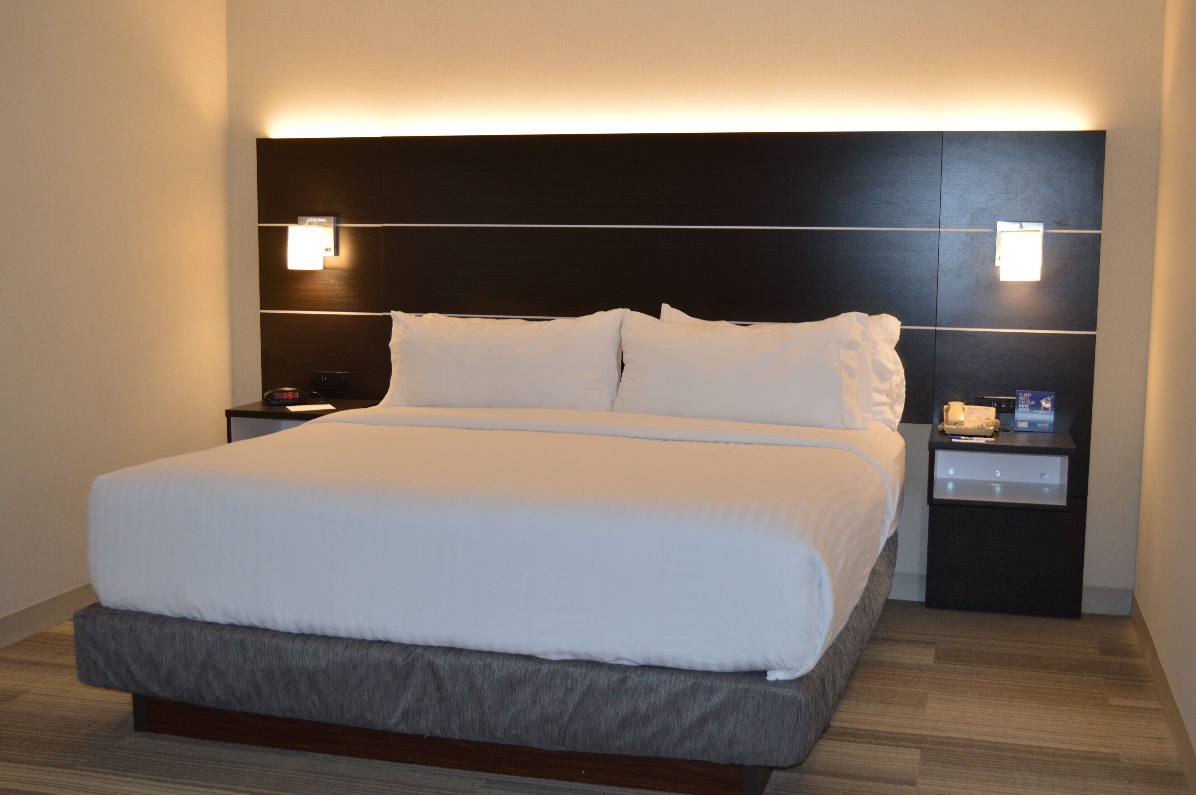 Una habitación de hotel moderna con una cama grande, un cabecero oscuro, mesitas de noche con lámparas y suelo de madera.