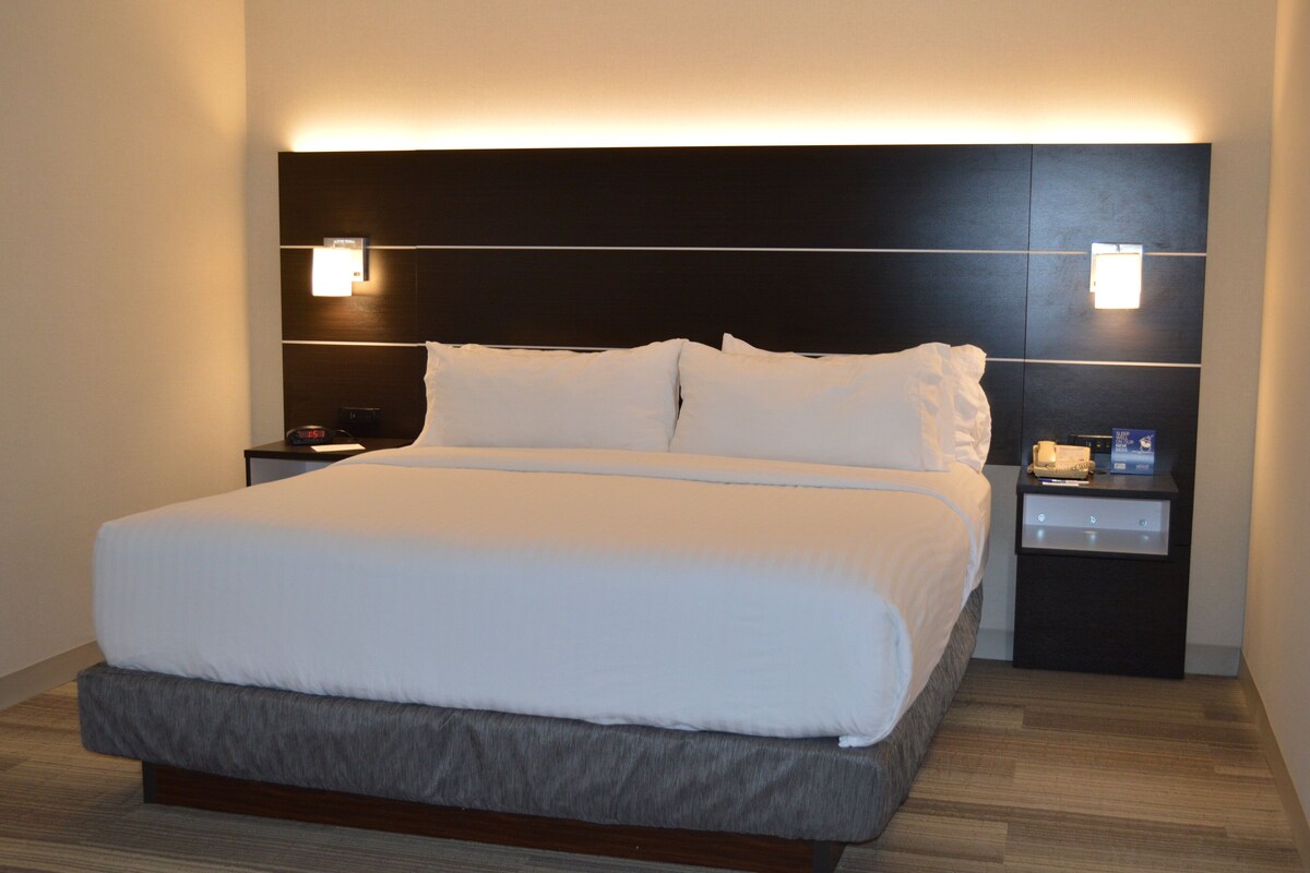 A modern hotel room with a large bed, a dark headboard, bedside tables with lamps, and a wooden floor.