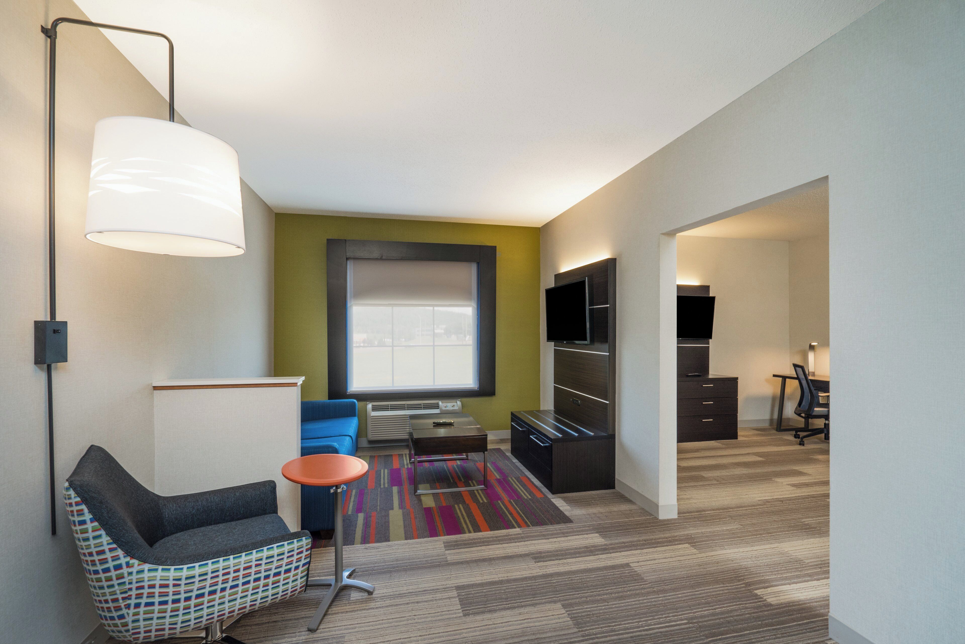 Una habitación de hotel moderna con sofá, una mesita, una alfombra colorida, un televisor y un escritorio con cajones.