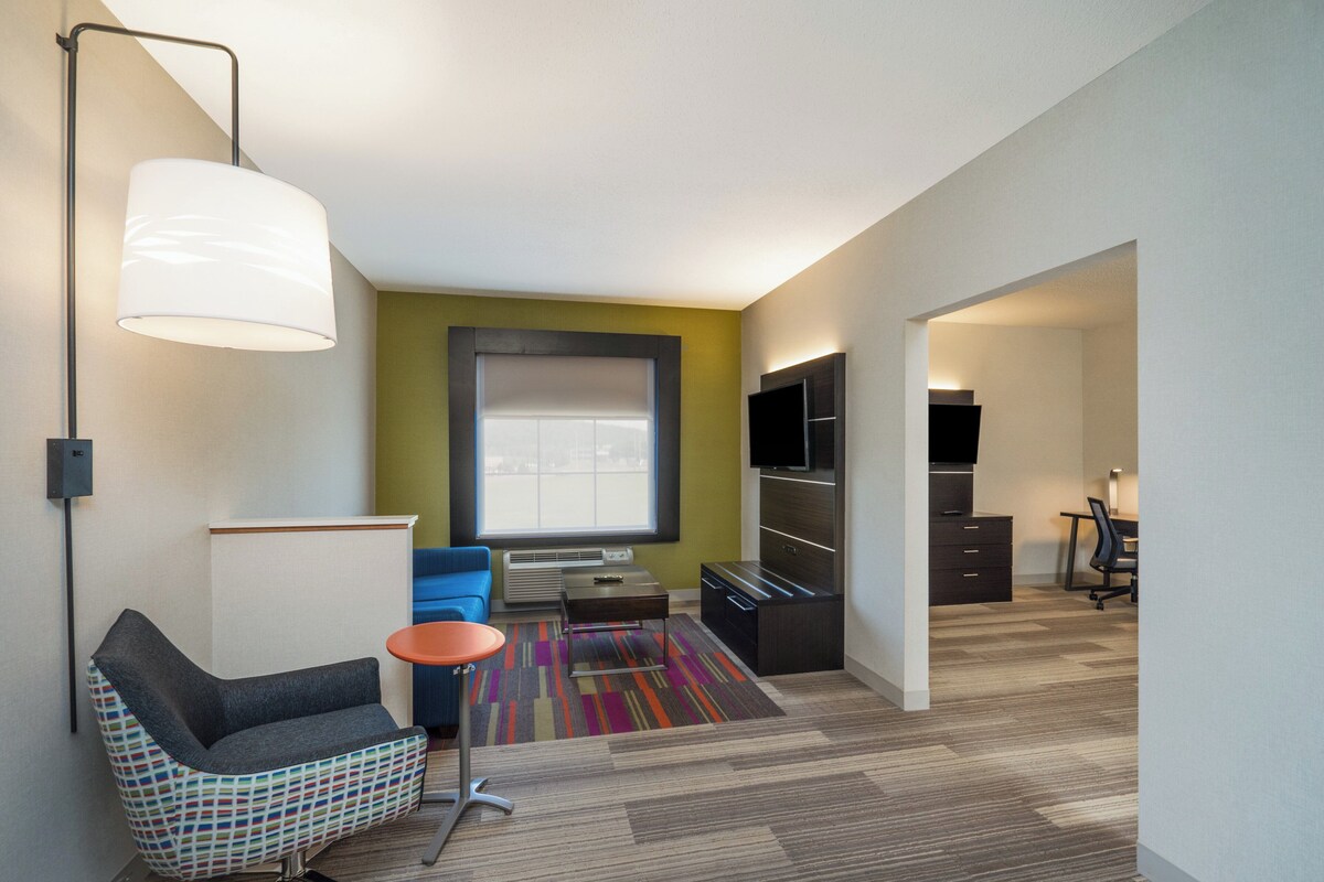 A modern hotel room with a large window, a sofa, a chair, a desk, and a wardrobe.