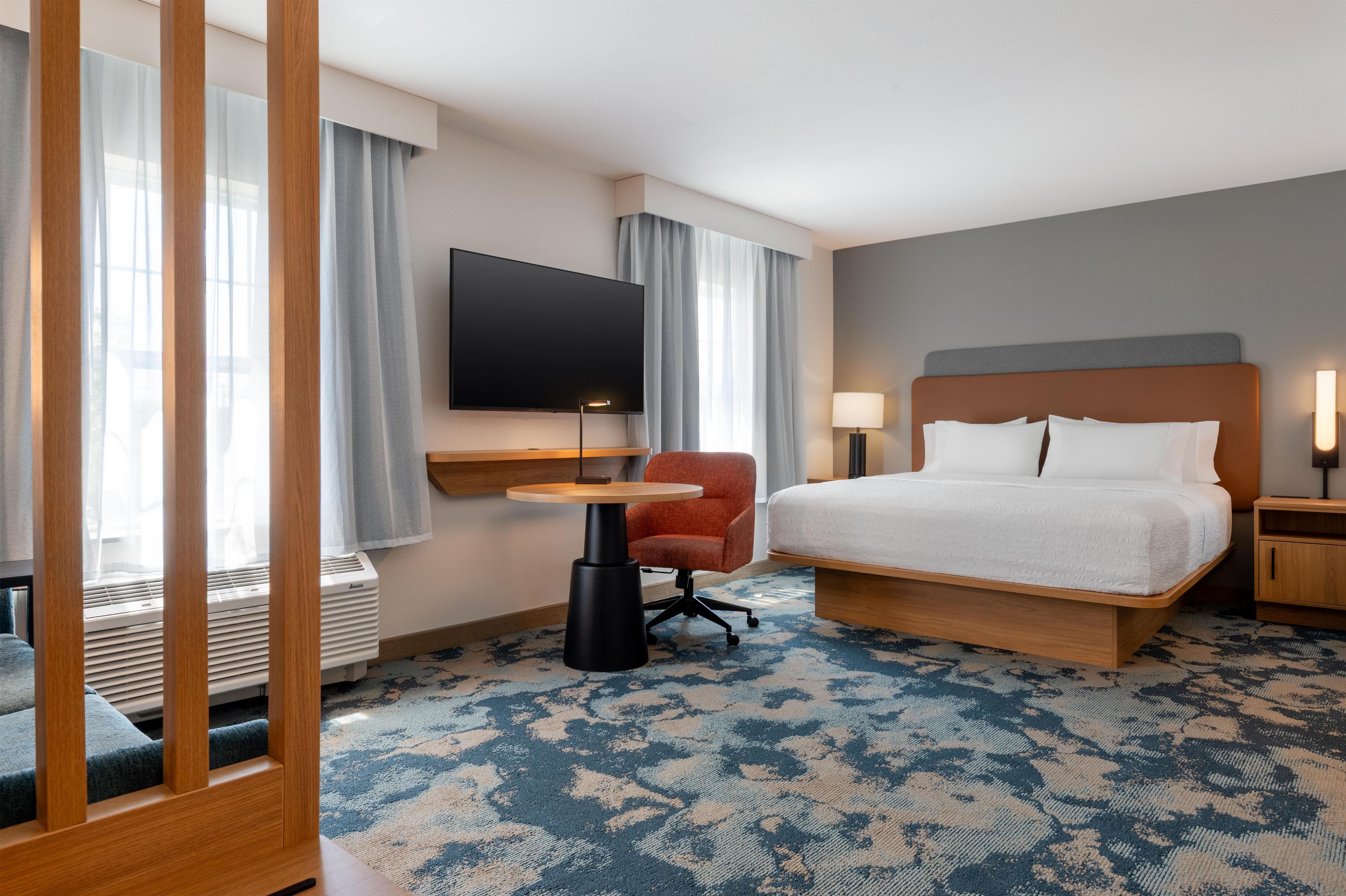 Habitación de hotel con cama, escritorio, silla, televisor y ventana con cortinas.