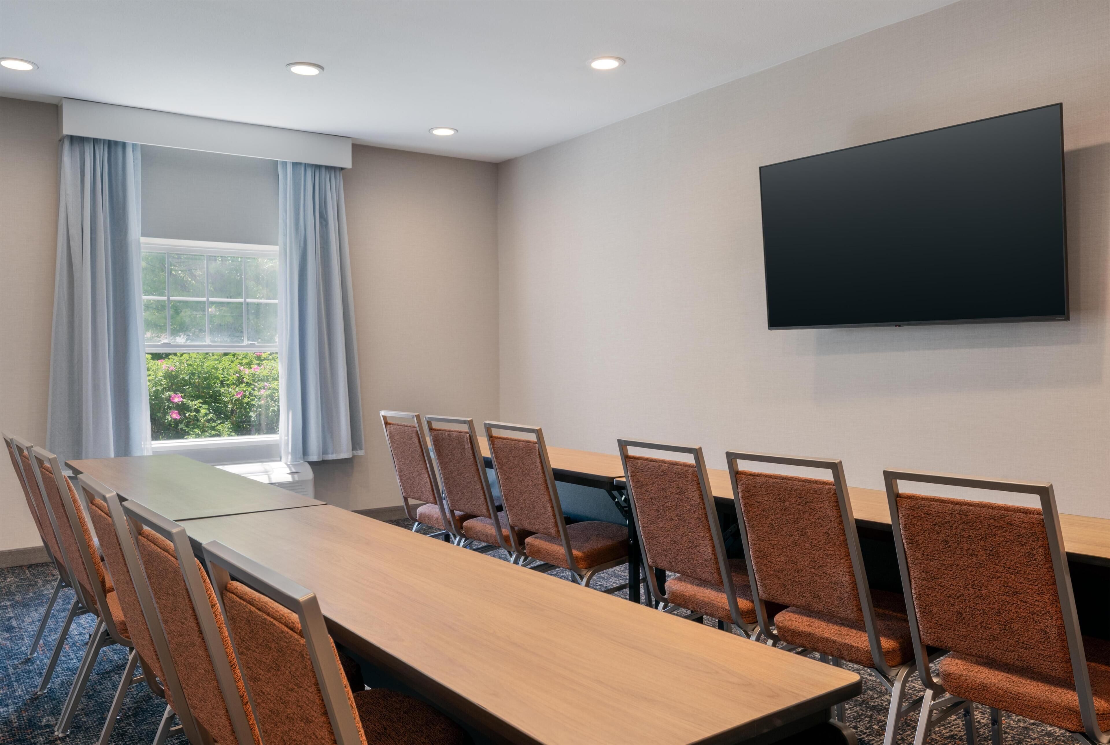 A conference room with a long table, six chairs, a flat-screen TV, and a window with curtains.