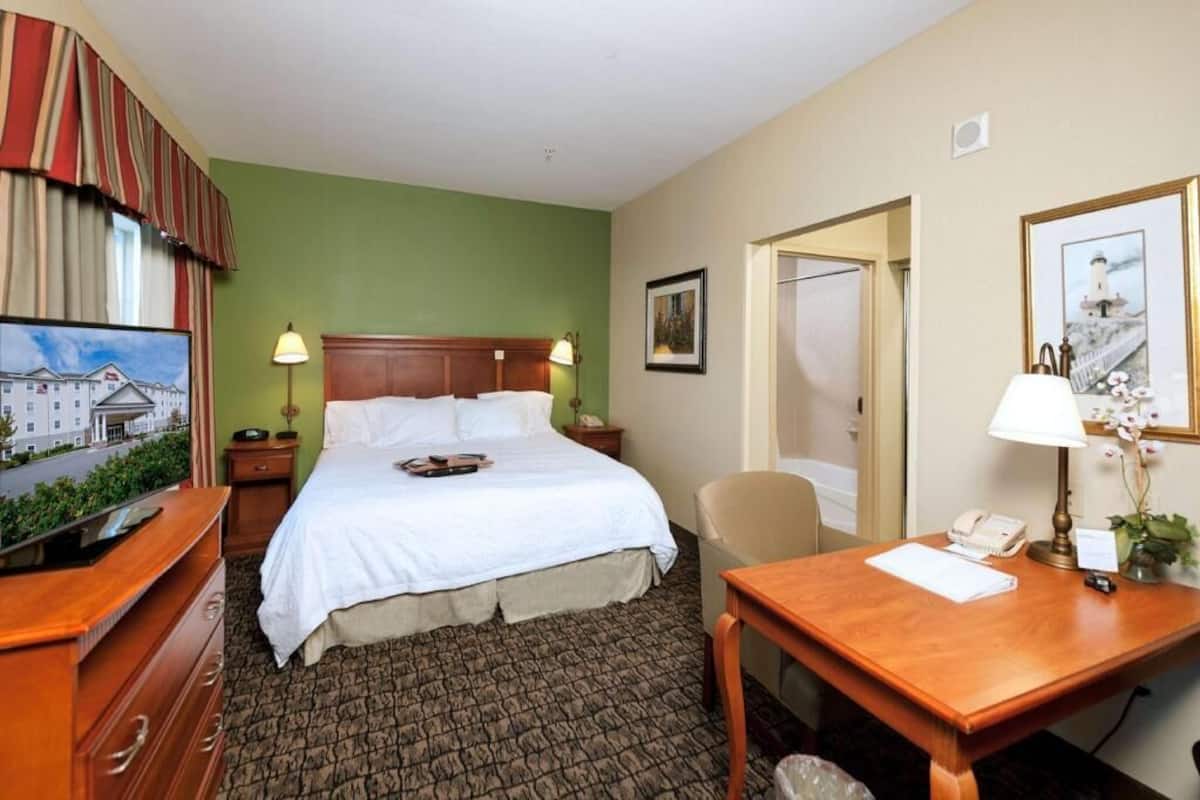 A hotel room with a bed, a desk, a chair, a lamp, a telephone, and a painting on the wall.
