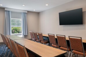 Een vergaderruimte met een lange tafel, zes stoelen, een flatscreen televisie en een raam met gordijnen.