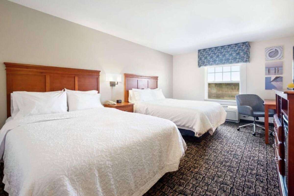 A hotel room with two beds, a desk, a chair, and a window with a view.