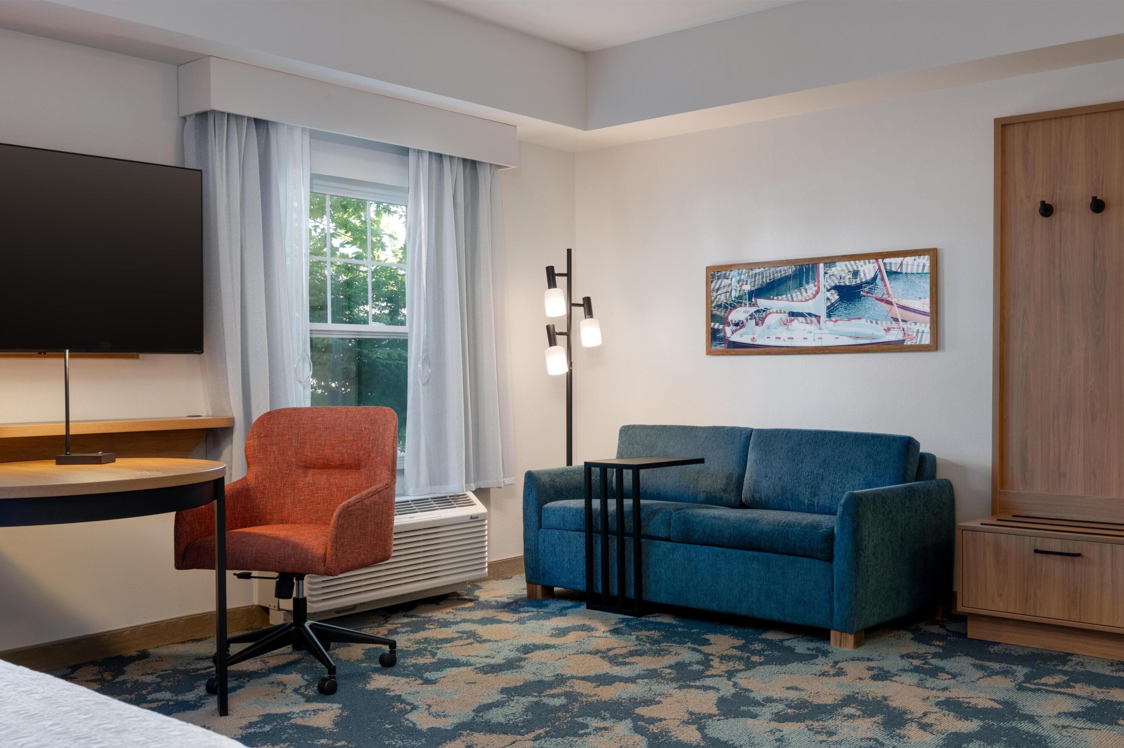 A hotel room with a desk, a blue sofa, a TV, a painting, and a wardrobe.