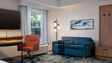 A hotel room with a desk, a blue sofa, a TV, a painting, and a wardrobe.