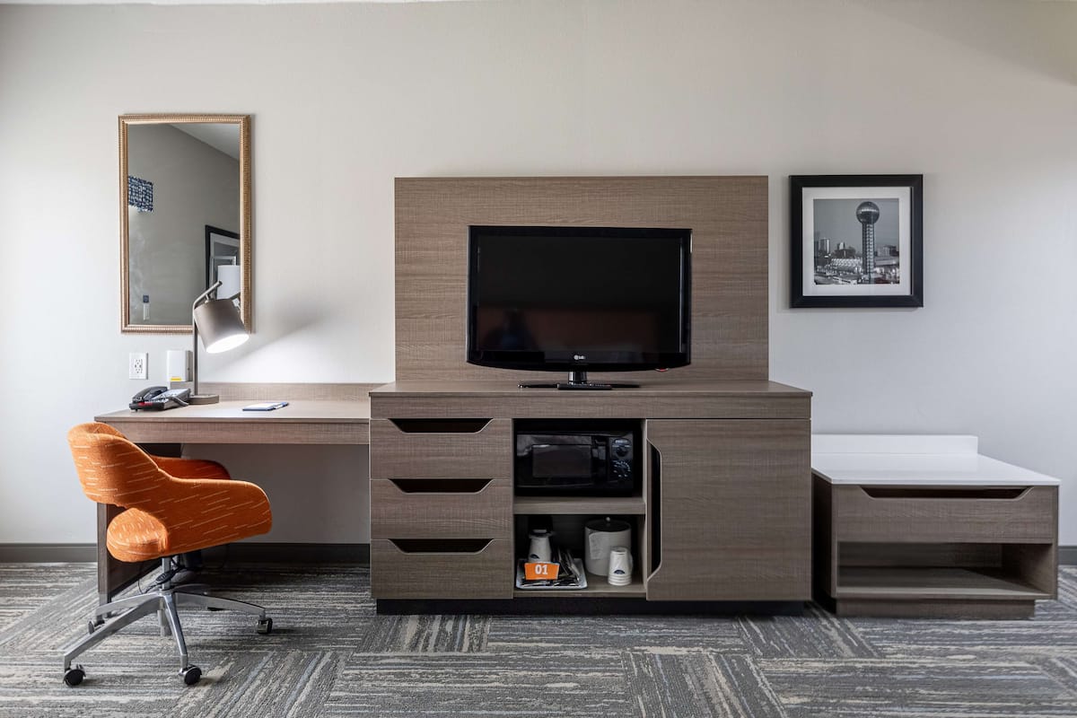 A hotel room with a wooden TV stand, a desk, a chair, a mirror, and a framed picture on the wall.