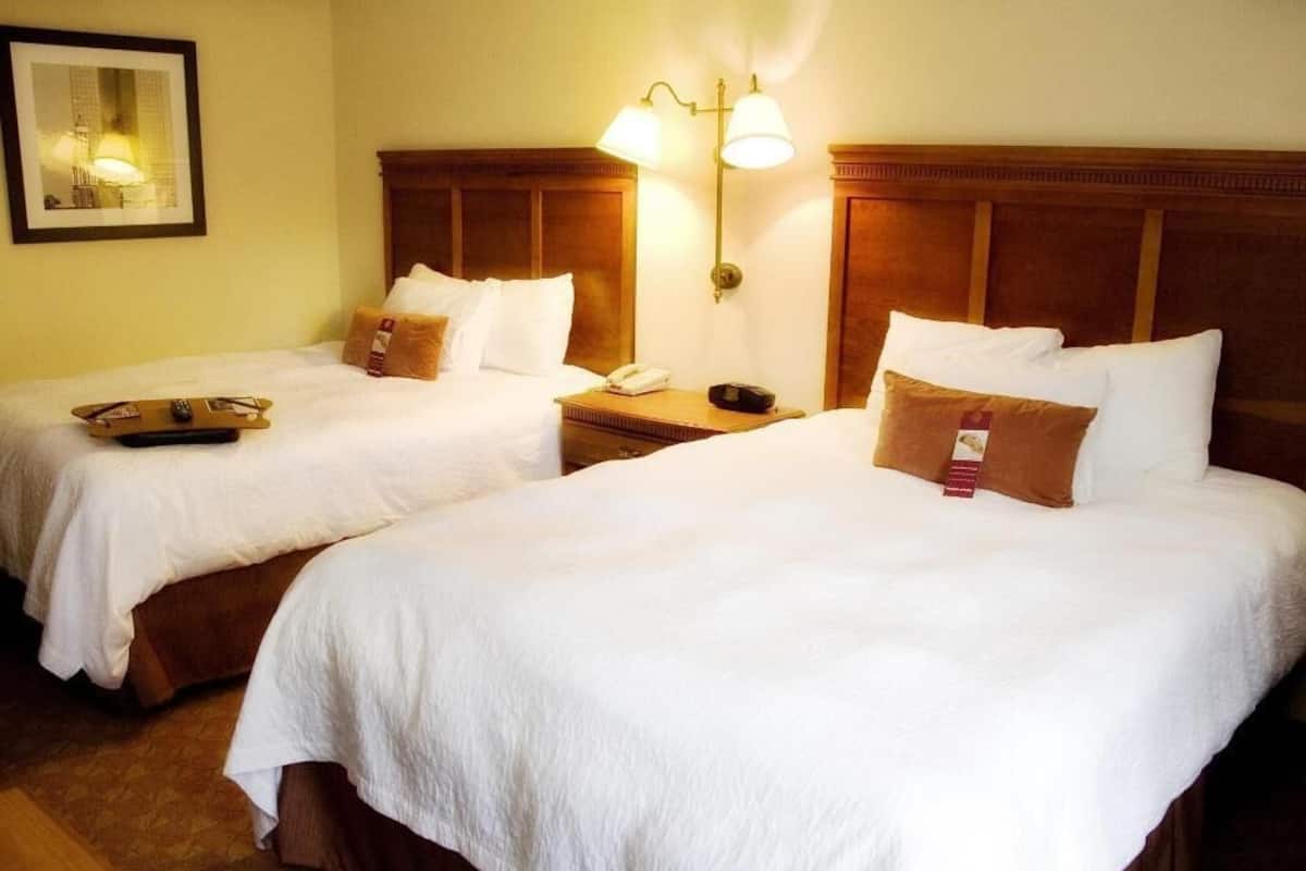 Ein Hotelzimmer mit zwei Betten, jeweils mit weißer Bettwäsche und braunen Kopfteilen. An einem Bett befindet sich ein Nachttisch mit einer Lampe und einem Telefon.