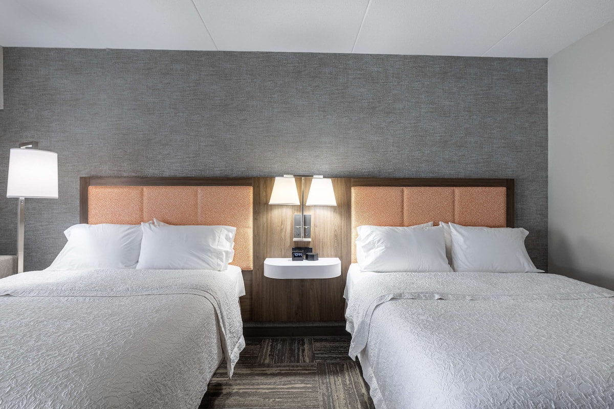 A hotel room with two beds, a wooden headboard, a built-in sink, and a nightstand with a lamp.