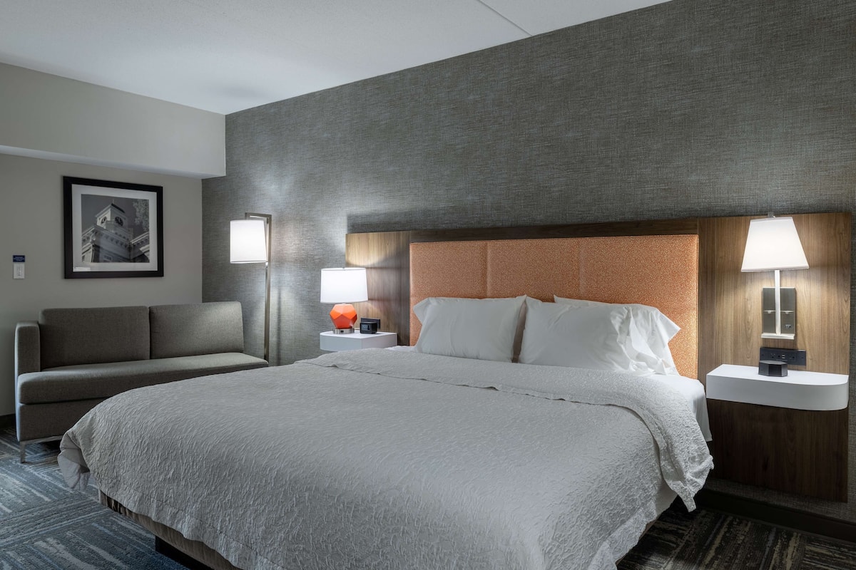 A hotel room with a large bed, two bedside tables with lamps, a sofa, and a framed picture on the wall.