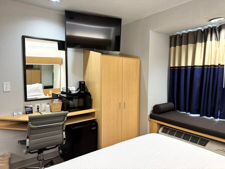 Une chambre d’hôtel comprenant un lit, un bureau avec une chaise, un four à micro-ondes, un petit réfrigérateur, un téléviseur fixé au mur et une fenêtre avec des rideaux.