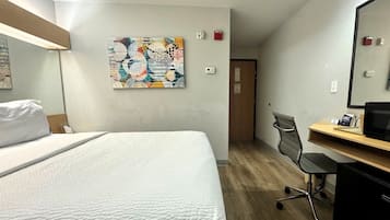 Ein Hotelzimmer mit einem Bett, einem Schreibtisch, einem Stuhl, einer Schreibtischlampe, einem Computer, einem Wandbild und einer Tür.