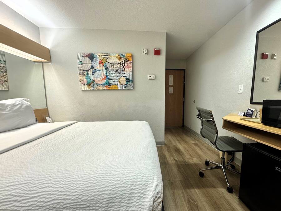 Ein Hotelzimmer mit einem Bett, einem Schreibtisch, einem Stuhl, einer Schreibtischlampe, einem Computer, einem Wandbild und einer Tür.