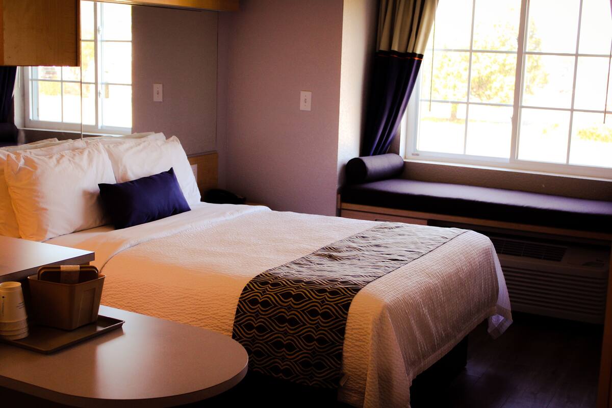 A hotel room with a bed, a bedside table with a cup and a tray, a window with curtains, and a built-in bench.