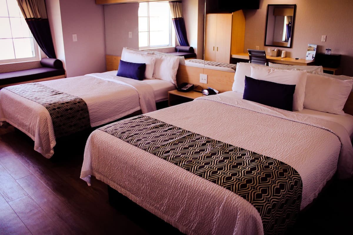 A hotel room with two beds, a nightstand, a desk, a chair, a mirror, and a window with curtains.