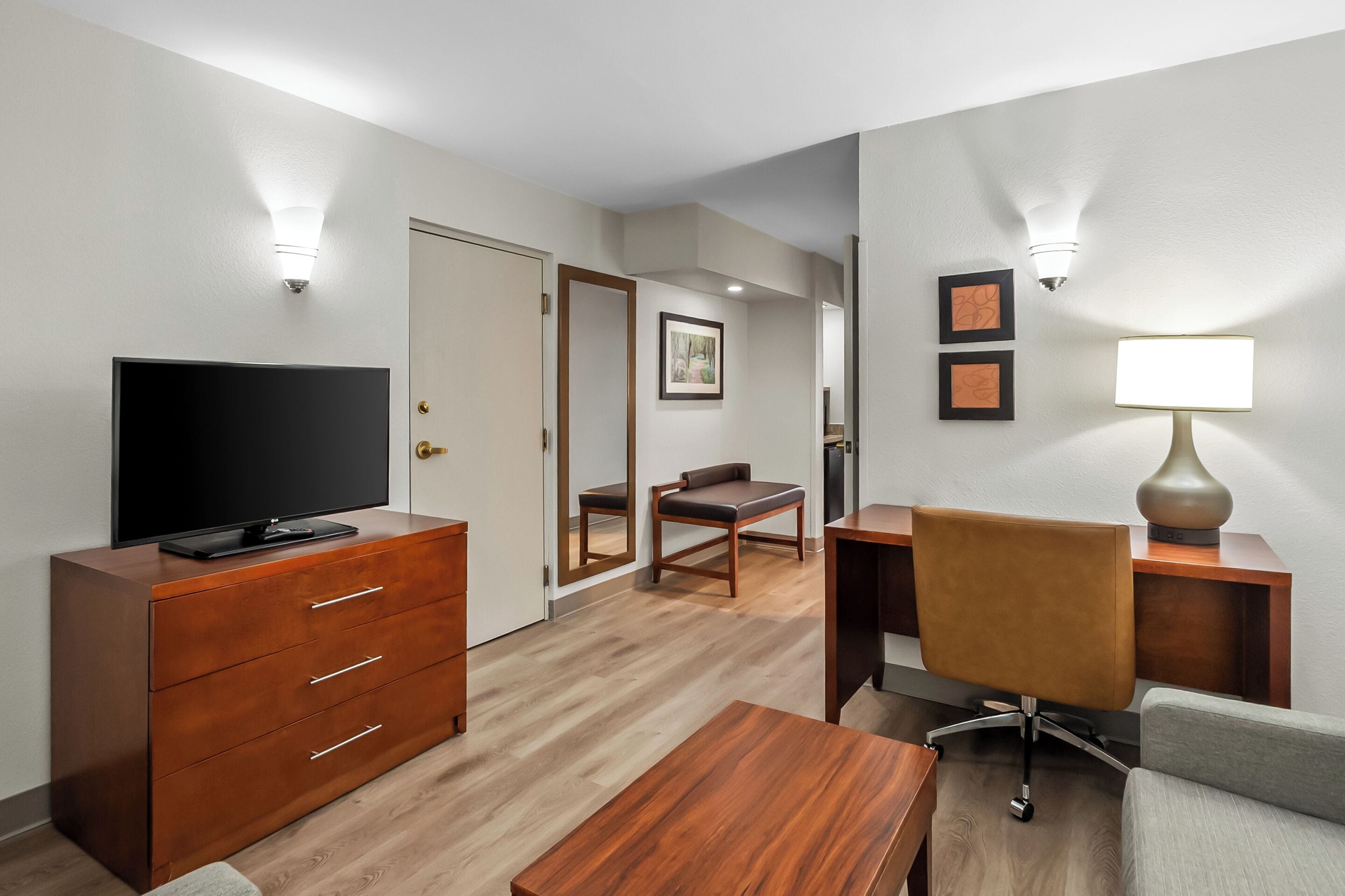 Habitación de hotel con televisión sobre un aparador de madera, un escritorio con silla, una lámpara y un espejo.