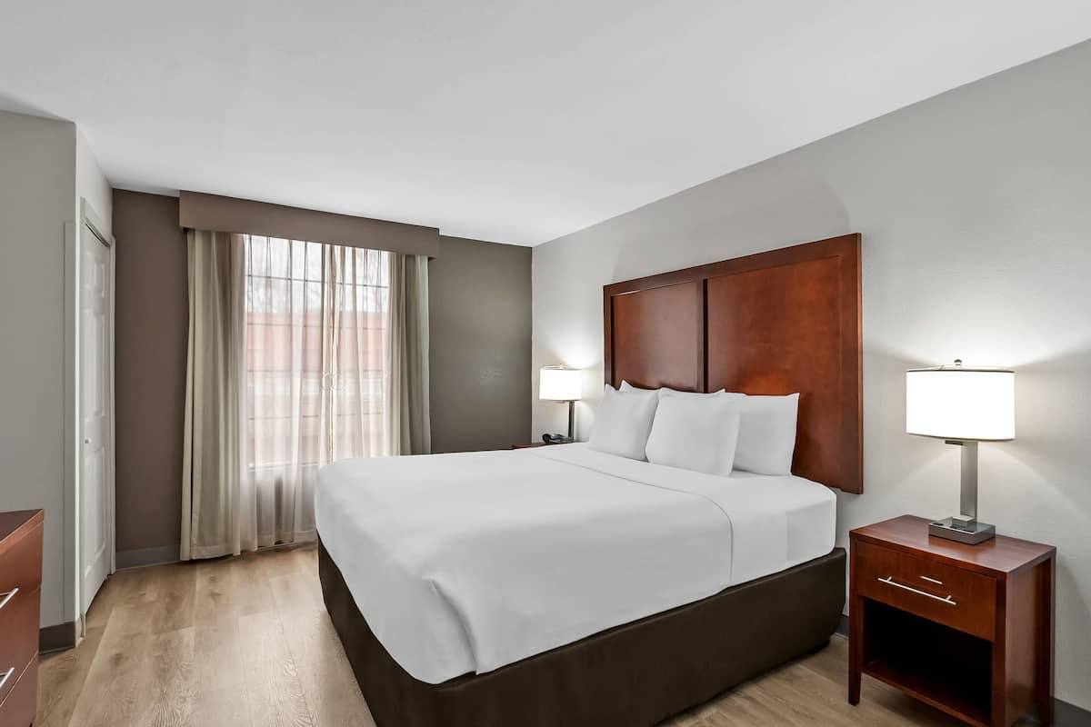 Een hotelkamer met een groot bed, twee nachtkastjes met lampen, een houten hoofdeinde en een raam met gordijnen.