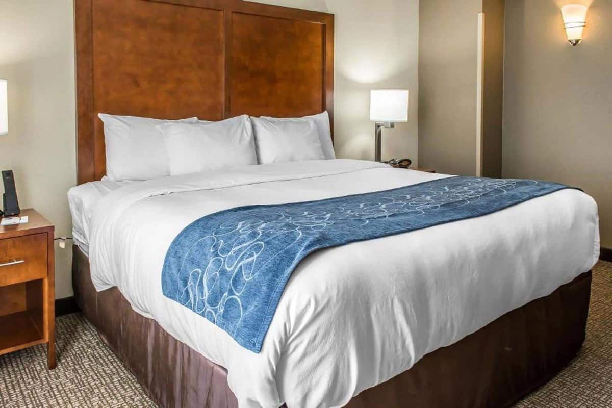 Een hotelkamer met een groot bed, een houten hoofdeinde, een nachtkastje met een lamp en een wandlamp.