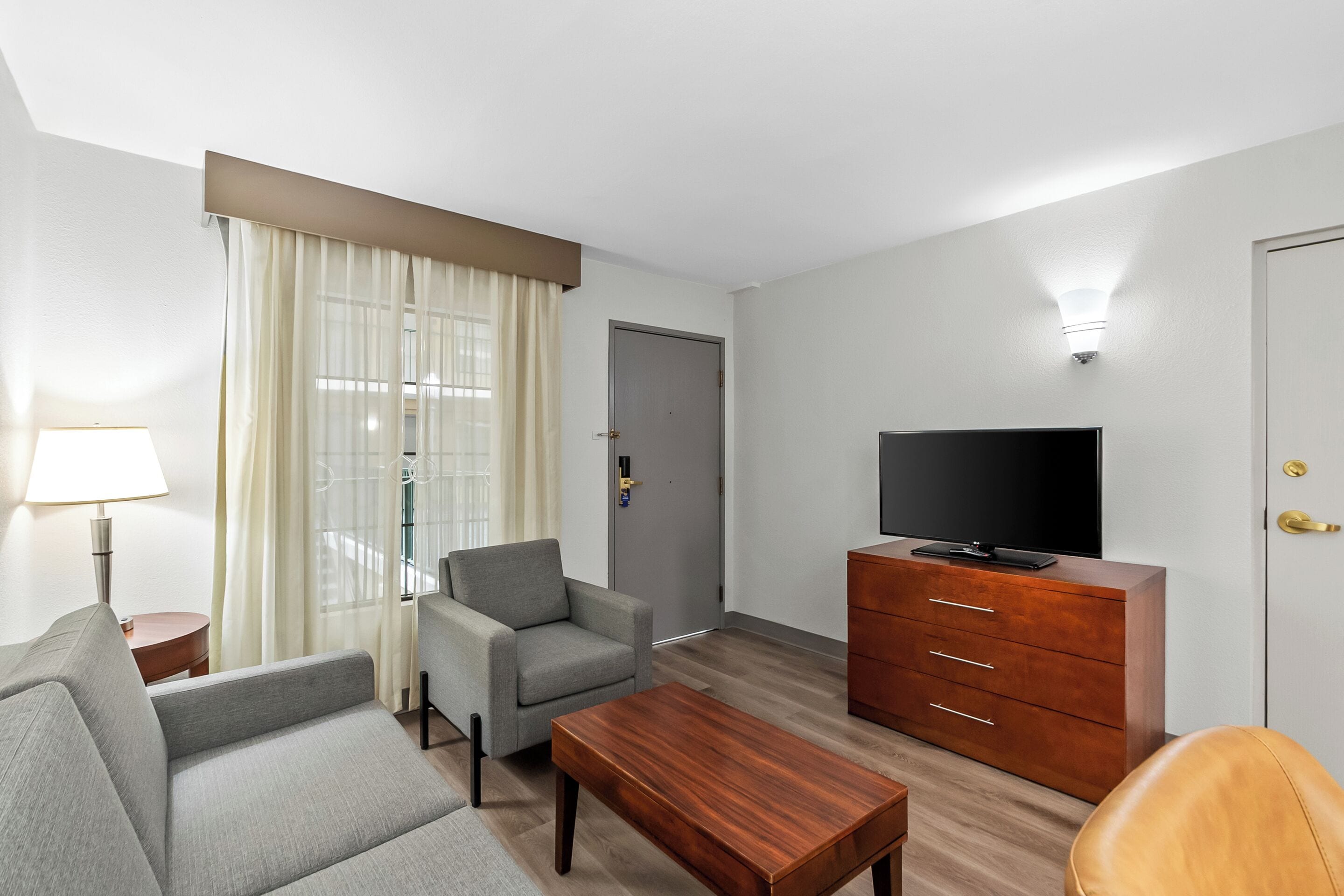 Ein Hotelzimmer mit einer Couch, einem Sessel, einem Holztisch, einem Fernsehmöbel und einer Lampe.
