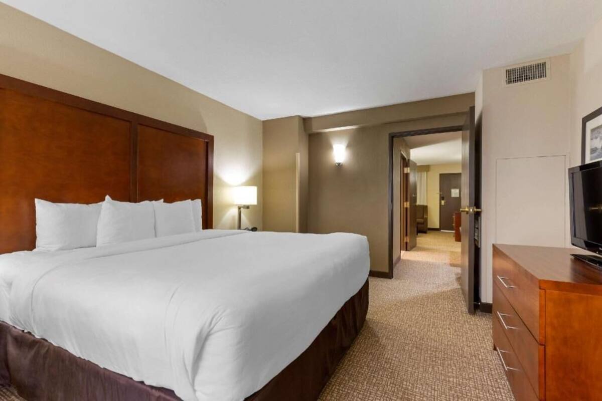 Een hotelkamer met een groot bed, een houten hoofdbord, een bureau met een televisie en een gang naar een andere kamer.