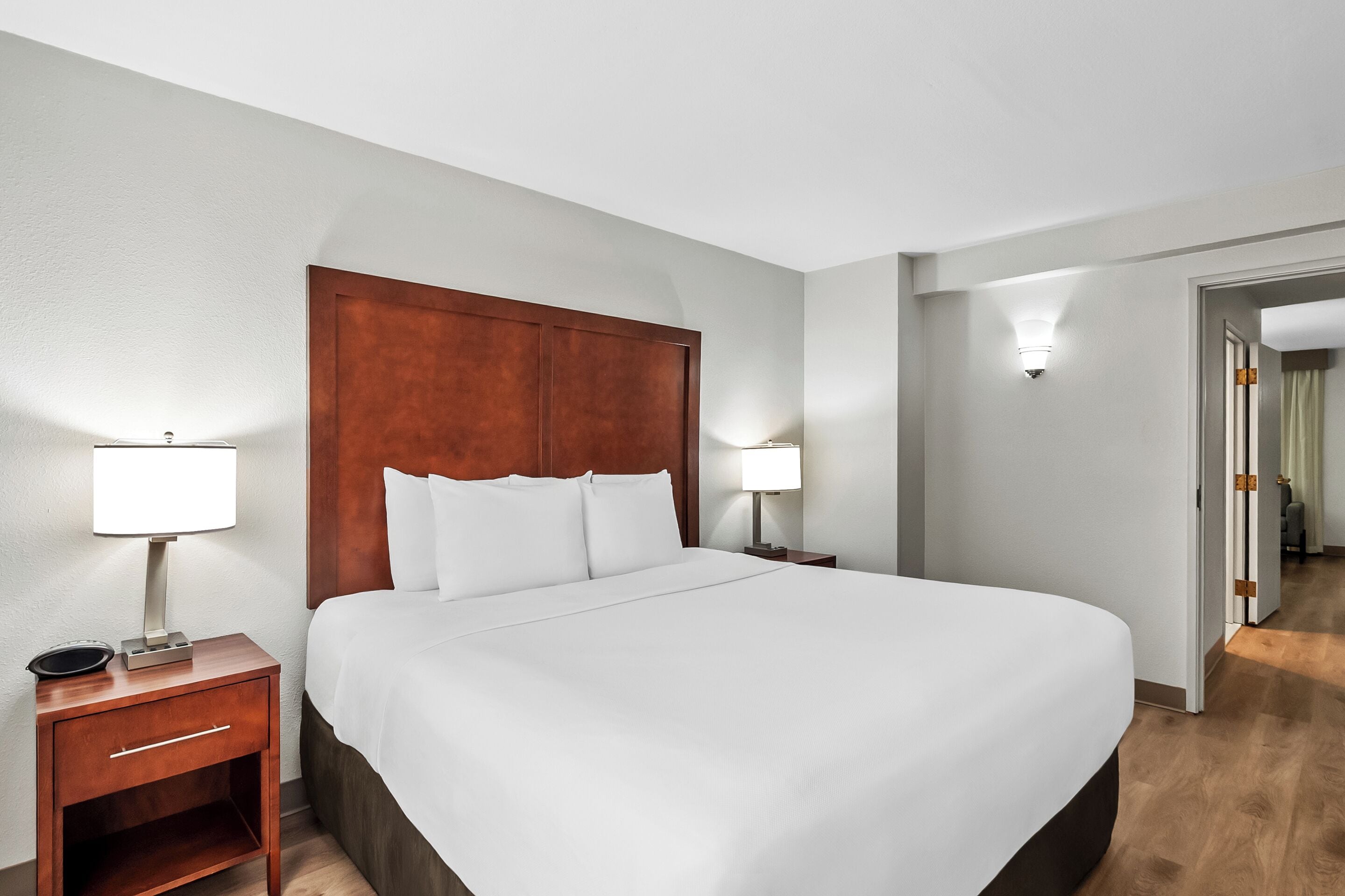 A hotel room with a large bed, two bedside tables with lamps, a wooden headboard, and a door leading to another room.