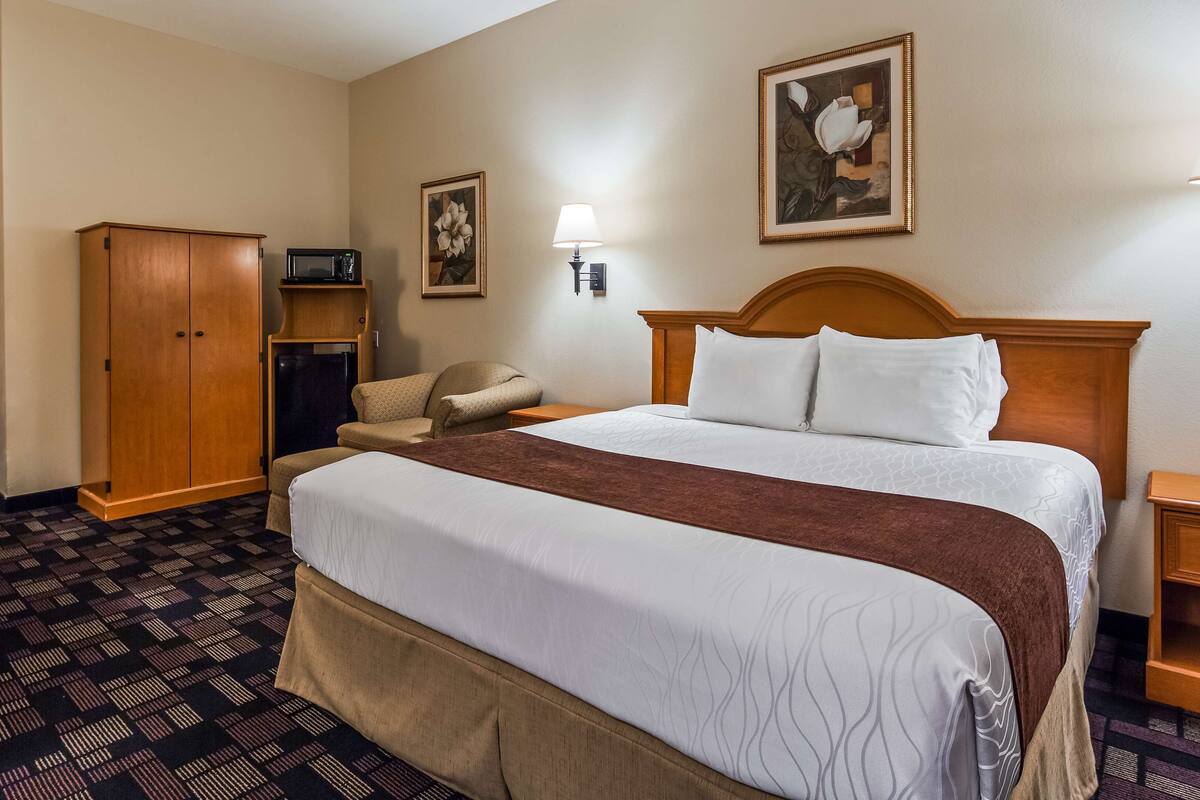 A hotel room with a large bed, a desk with a chair, a wardrobe, a microwave, and a painting on the wall.