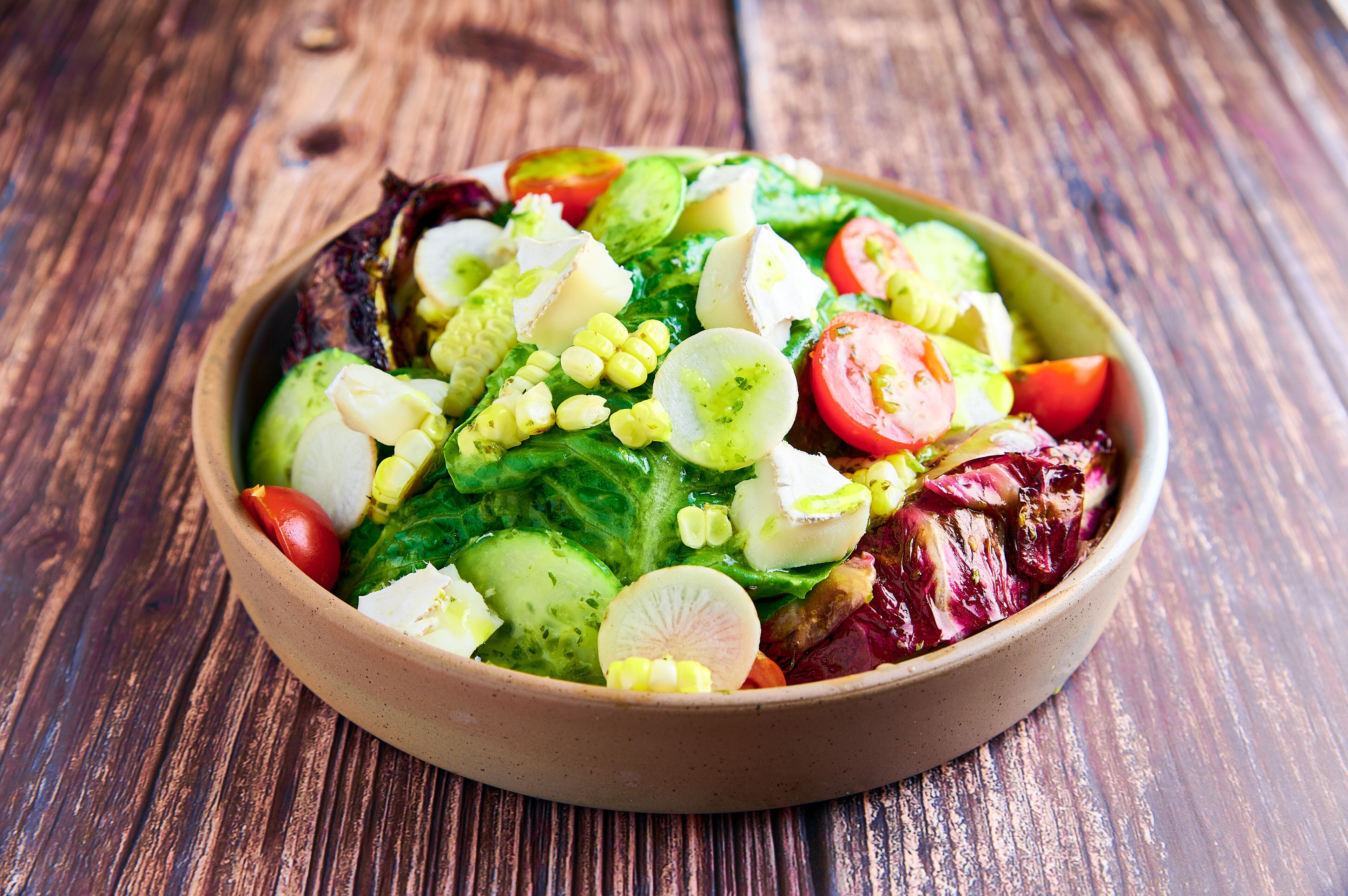 A bowl of salad with various vegetables and cheese on a wooden table.