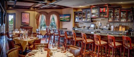 Un restaurante con mesas redondas, sillas de madera, una zona de bar con vitrinas de vidrio y vistas amplias a través de grandes ventanales.