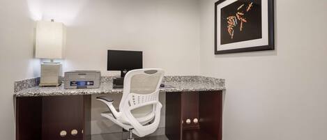 A modern office desk with a computer, printer, and lamp. There is a framed artwork on the wall and a white office chair in front of the desk.