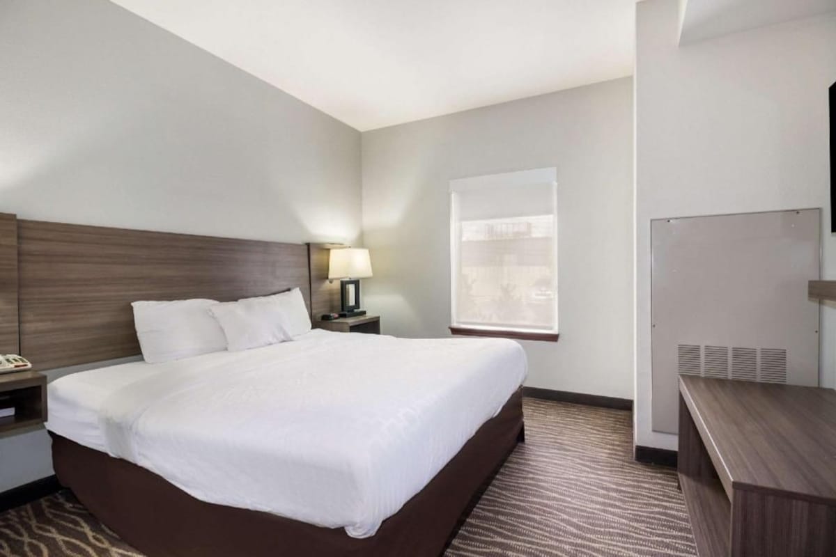 A hotel room with a large bed, a bedside table with a lamp, a desk, and a window with blinds.