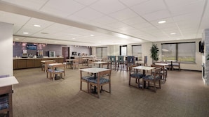 A spacious dining area with multiple tables and chairs, a kitchen area in the background, and large windows providing natural light.