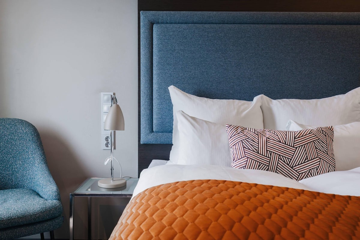 Habitación de hotel con una cama grande, cabecera azul, mesita de noche con lámpara y una silla azul.
