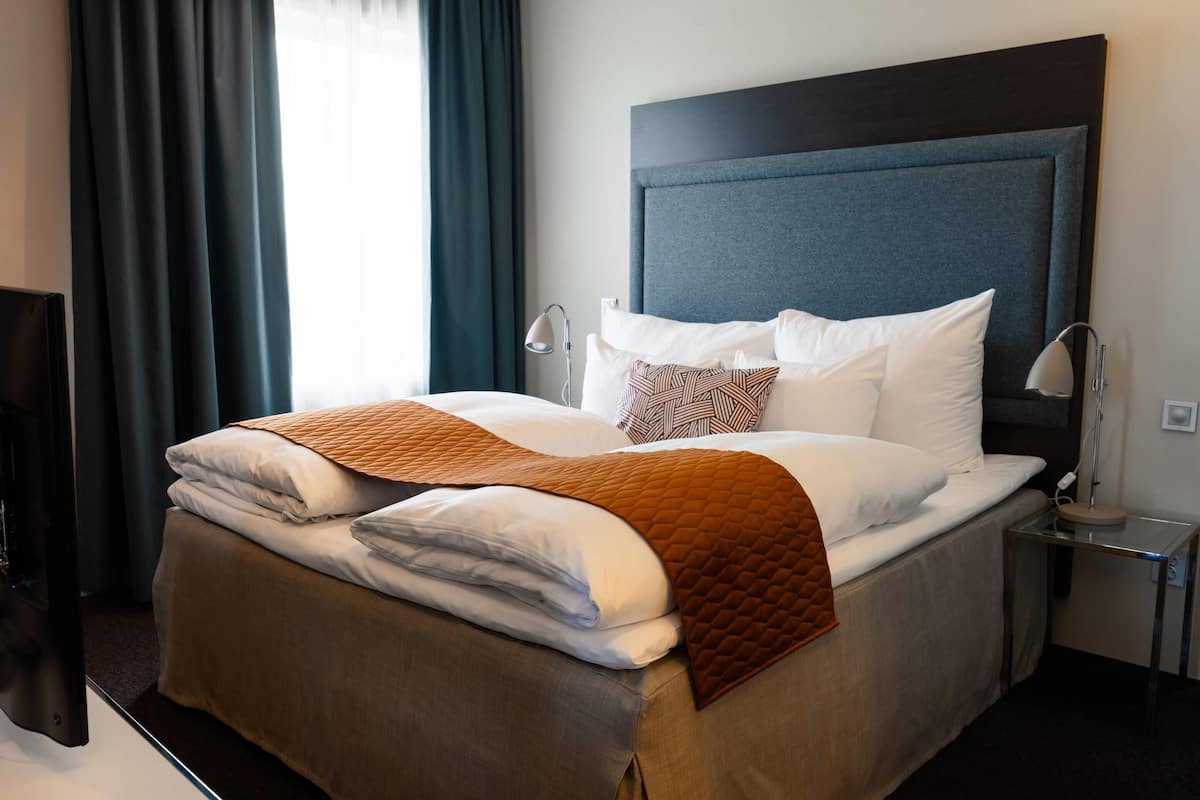 Una cama bien hecha con colcha marrón, almohadas blancas y cabecera oscura en una habitación de hotel.
