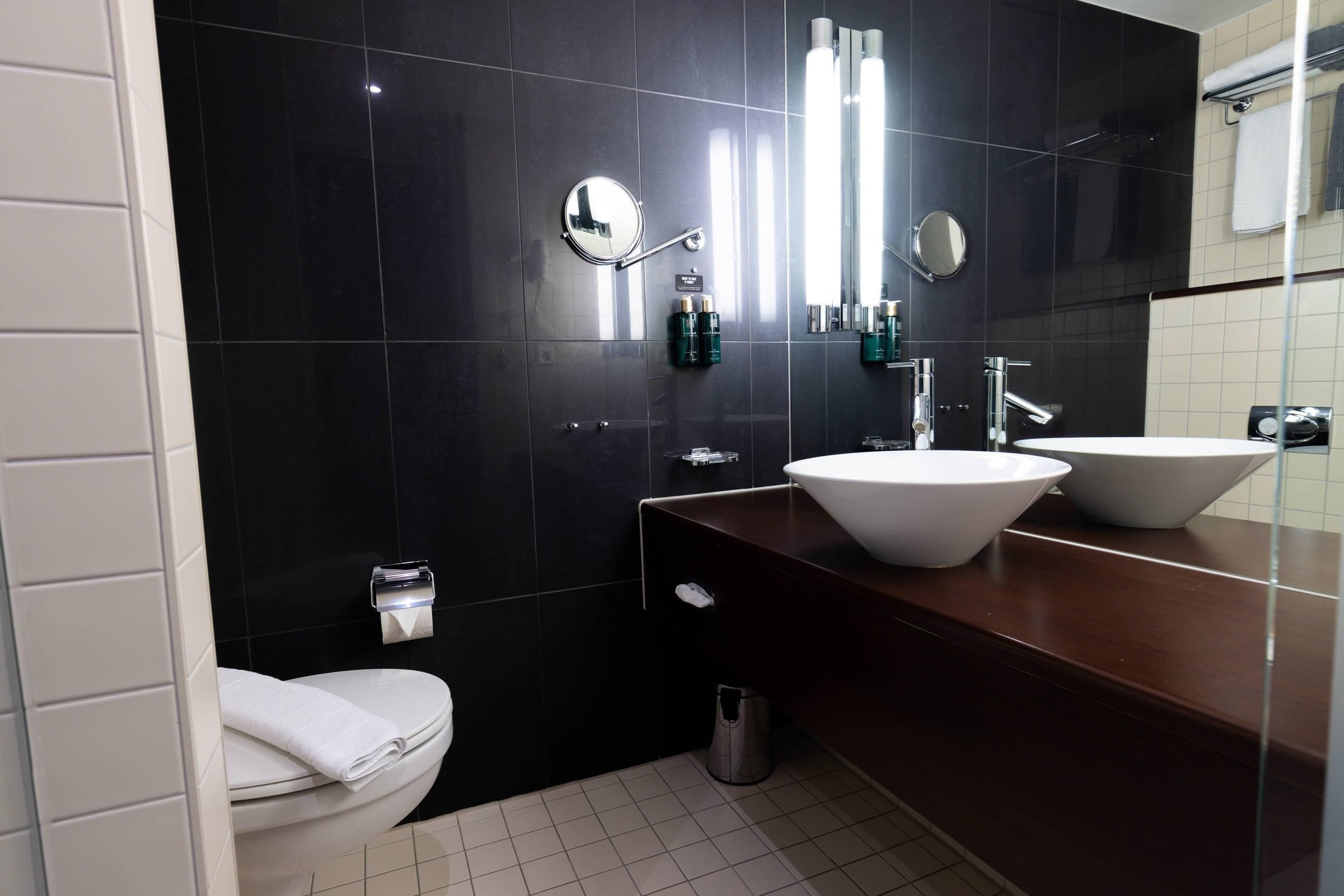 Un baño moderno con pared de azulejos negros, dos lavamanos blancos, un espejo y un inodoro.