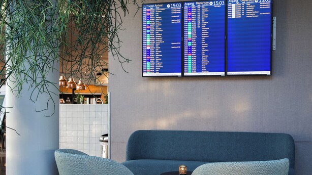 Un espace détente avec un canapé bleu et une petite table, un panneau d’affichage numérique indiquant les informations sur les vols, et une plante.