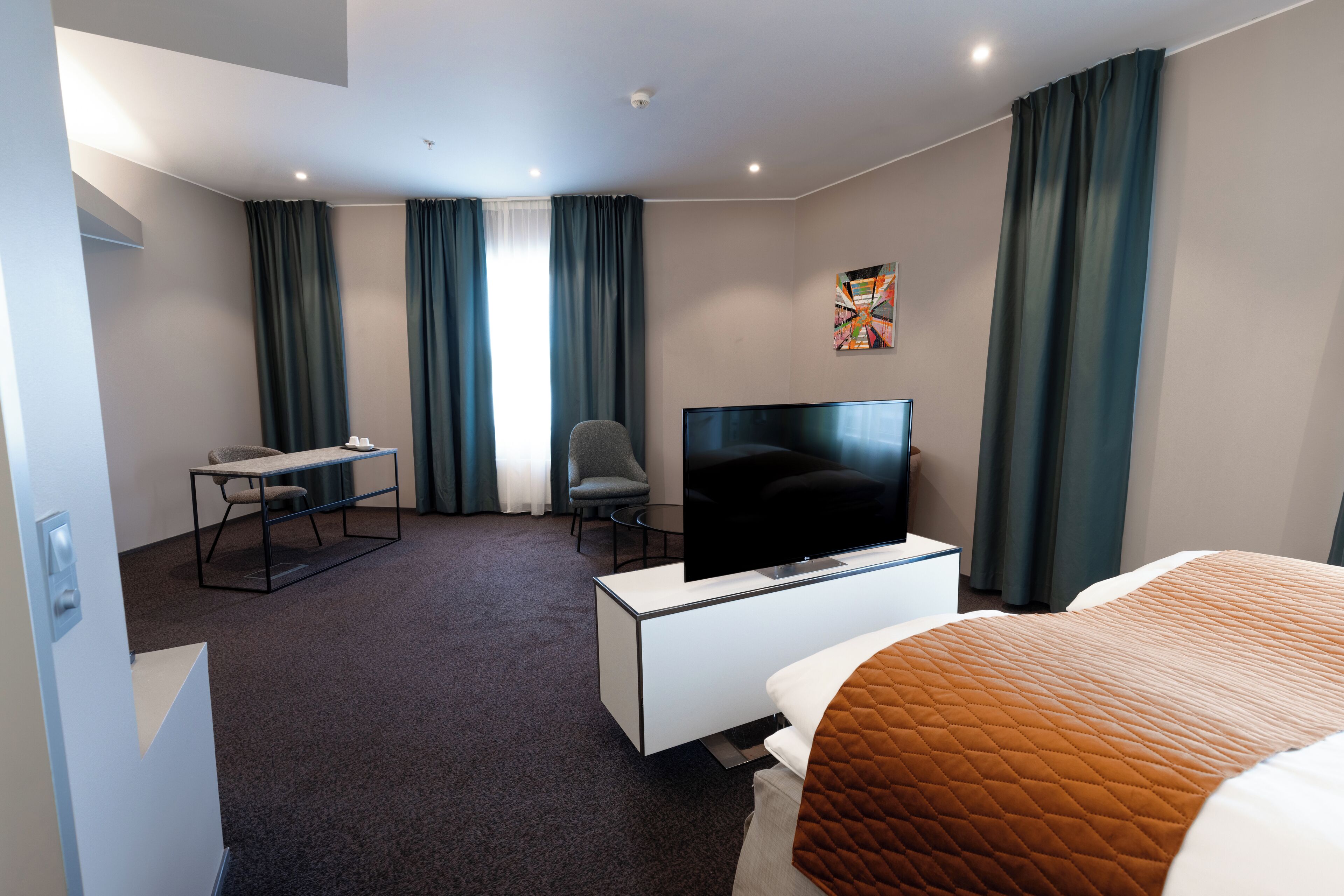 A hotel room with a bed, a desk with a chair, a television, and a painting on the wall.