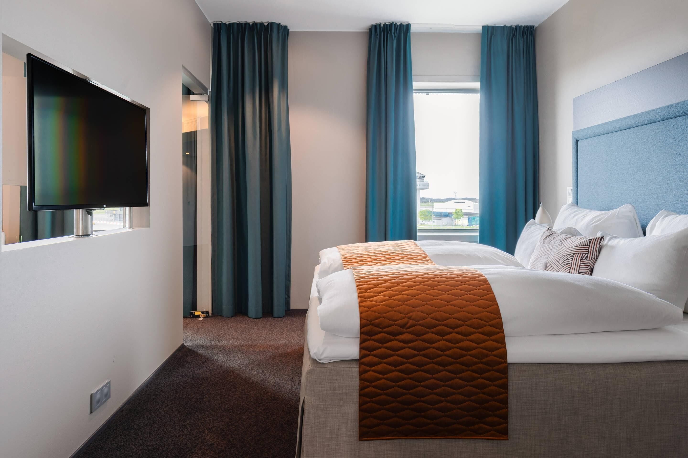 A hotel room with a bed, a flat-screen TV, and a view of the outside through large windows.