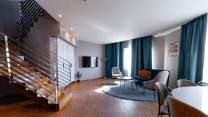 Una sala de estar moderna con una escalera de madera, un televisor de pantalla plana, un comedor con mesa y sillas, y un ventanal con cortinas.