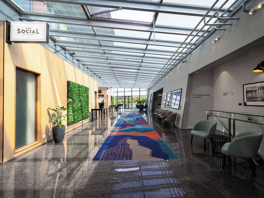A modern hotel corridor with a glass ceiling, a reception desk, seating area, and a colorful carpet.