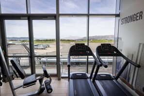 Ein Fitnessstudio mit Laufbändern und Kraftgeräten, große Fenster mit Blick auf einen Flughafen und Berge.