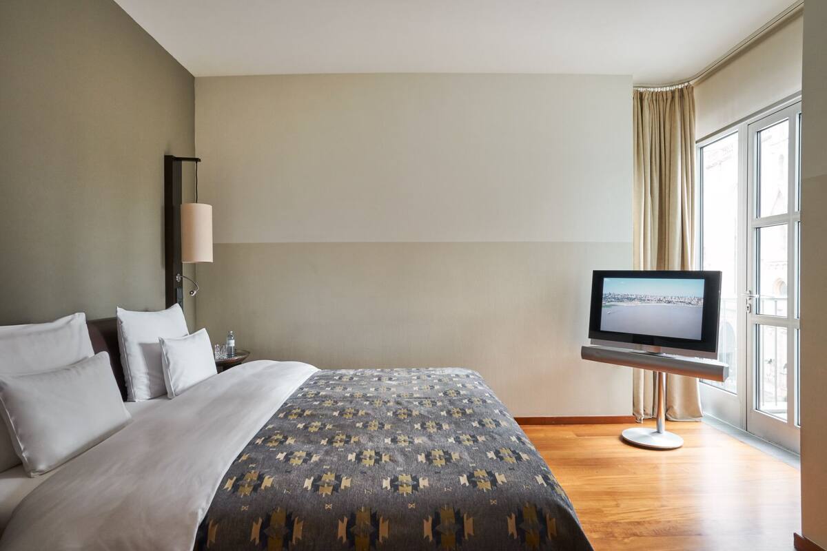 A hotel room with a bed, a television on a stand, a nightstand, and a window with curtains.