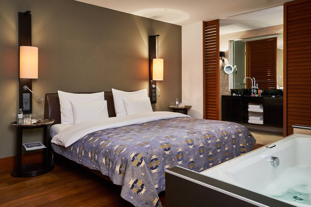 A hotel room with a large bed, a bedside table with a lamp, a bathroom with a sink and mirror, and a bathtub.