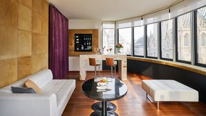 A modern living room with a white sofa, a round table with a bowl of fruit, and a view of a cityscape through large windows. - DO & CO Hotel Vienna (Vienna)